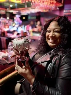 A woman smiling and holding a camera in a vibrant bar or nightclub setting with colorful lights.