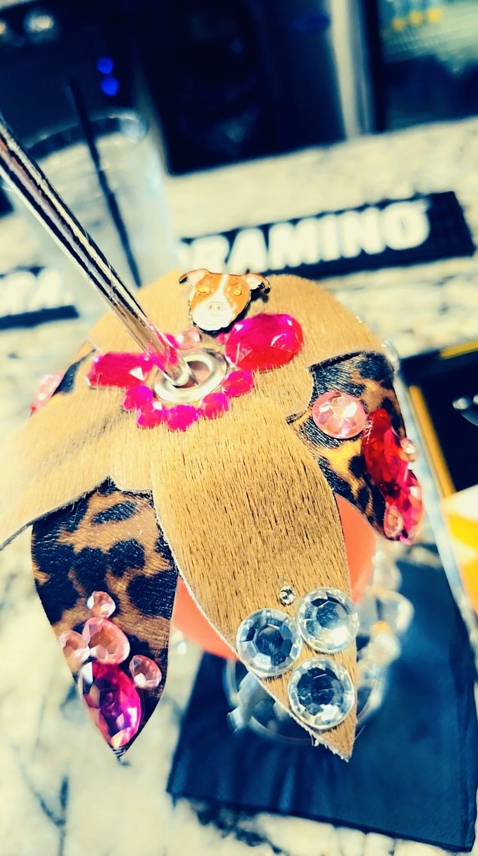 Close-up of a decorative item resembling a dog's face, made of wood and decorated with pink rhinestones and leopard-print fabric, with a glass straw inserted into the top.