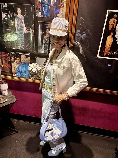 Woman wearing a white jacket, striped cap, and white pants, smiling and holding a blue floral purse, standing in front of framed photos on a burgundy wall.