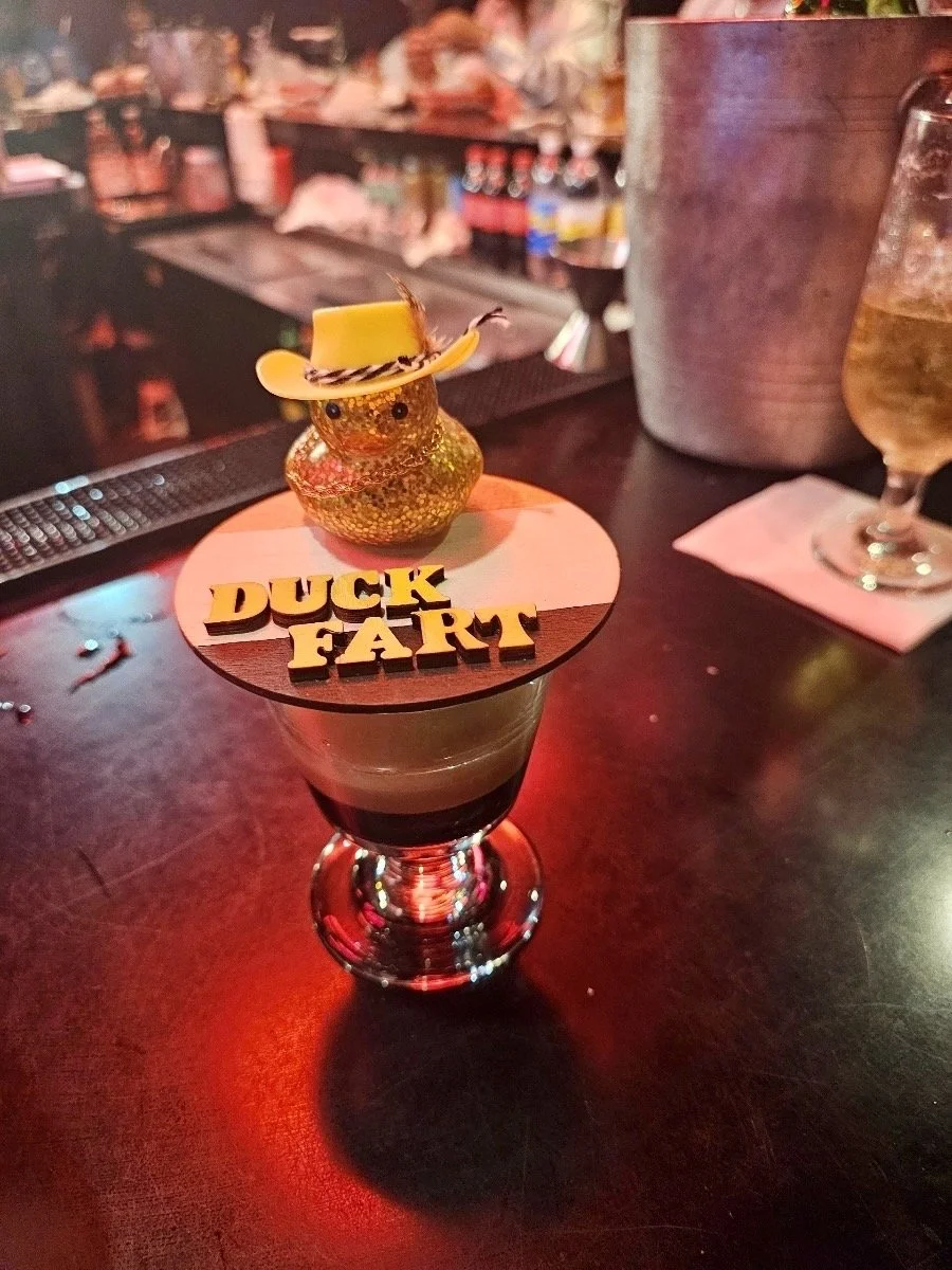 A dessert or drink in a tall glass topped with a decorative object that resembles a small chick wearing a yellow hat and a striped scarf, placed on a round chocolate wafer with yellow letters spelling 'DUCK FART'. The background shows a bar with bott