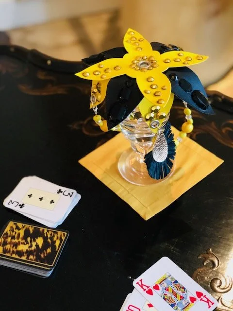 Decorative artificial flower arrangement with yellow and black flowers, adorned with rhinestones, placed in a glass on a wooden coaster on a black table, with a deck of playing cards nearby.