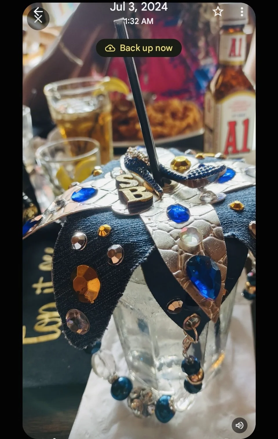 A decorated glass with a lid adorned with blue, gold, and clear rhinestones, and a black straw, on a table with drinks and food in the background.