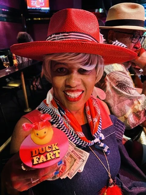A woman smiling and dressed in colorful clothing with a red hat, holding a bubble tea drink with a duck-shaped decoration labeled 'Duck Fart'. She is in a lively indoor setting with other people.