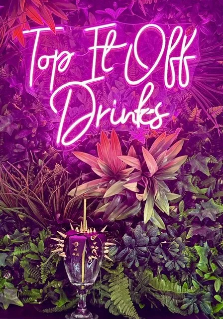 Pink neon sign stating "Tapa Fi Oka Drinks" surrounded by lush green and purple tropical plants, with a glass holding a purple dessert adorned with small toppings and a lit candle at the front.