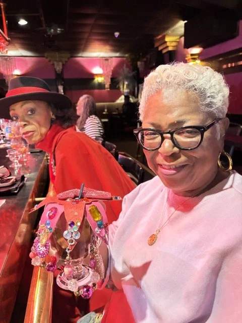 Two women sitting at a bar in a dimly lit, colorful nightclub or lounge. The woman in the foreground has gray curly hair, glasses, earrings, and a light pink top. The woman in the background is wearing a red hat and matching red outfit, with dark hai