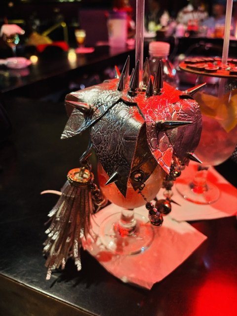A decorative cocktail glass with metallic studded leaves and keychain accessories, set on a bar counter with other drinks and blurred background.