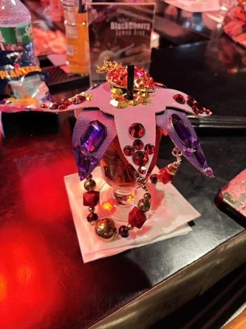 Decorative item resembling a pink and purple dragon with gems, beads, and feathers, placed on a table with a drink and other objects in the background.