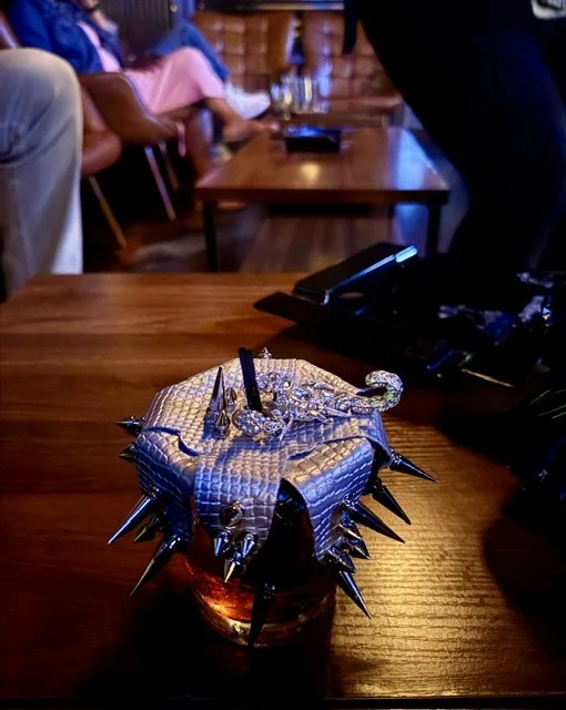 A glass with a black, spiked, armored lid resembling a dragon or monster head, made of textured material, sits on a wooden table.