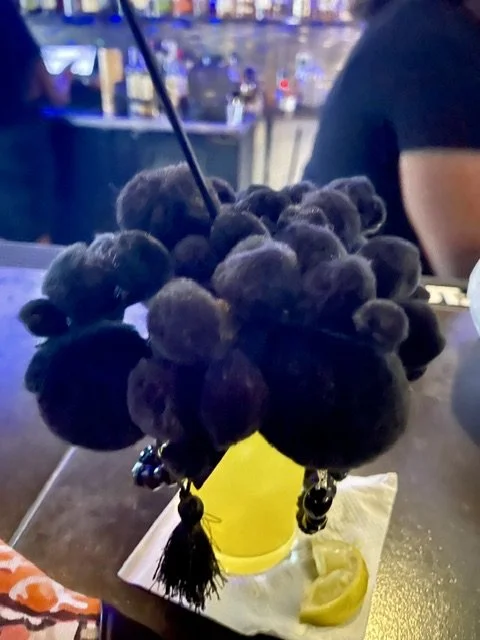 A plant with numerous round, dark purple to black clusters resembling berries or grapes, placed in a small yellow pot on a brown table at a restaurant or cafe.