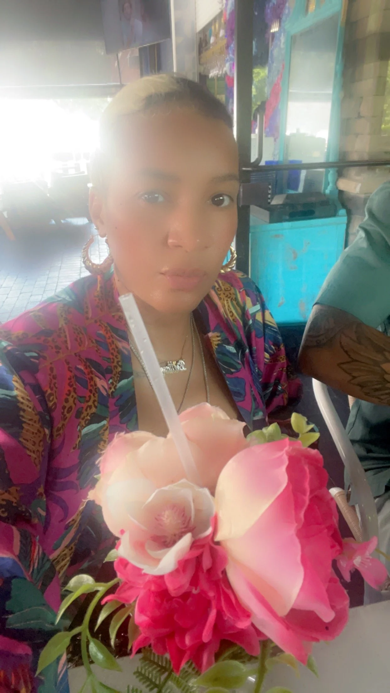 A woman with light brown skin and dark hair styled in a bun, wearing colorful clothing and large gold earrings, sitting at a table in a restaurant with a bouquet of pink and white flowers in the foreground, holding a drink with a straw.