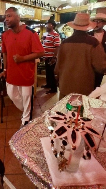 A birthday cake decorated with pink and black accents, a lit birthday candle, and candy or decorative beads, on a sparkly tablecloth. In the background, people are socializing in a restaurant or bar setting, some wearing hats and casual attire.