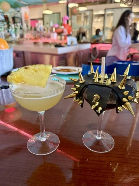 Two cocktails on a bar counter, one with a pineapple slice garnish and the other with a black leather band decorated with gold spikes, in a lively bar or restaurant setting.