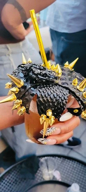 Person holding a cupcake decorated as a black crocodile with gold accents, including gold spikes and a yellow candle.