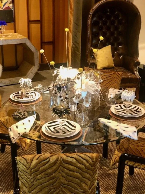 A dining table set with black and white patterned plates, silverware, and decorative glassware, with a floral centerpiece, two glasses filled with beverages, and a stylish chair with a tiger-striped cushion in the background.