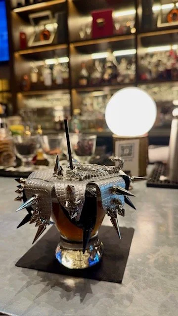 A cocktail in a unique, spiked silver cup with two black straws, on a black napkin at a bar counter, with illuminated shelves and a glowing round lamp in the background.