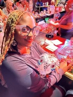 A woman with blonde braided hair and glasses sitting at a bar counter, holding shot glasses of multiple liquors, with people in the background.