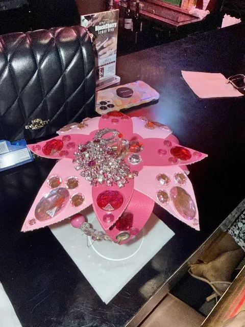 Pink paper craft in the shape of a flower with various rhinestones, beads, and decorative items on top, placed on a table in a dark room.