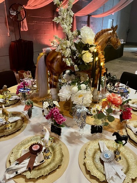 Decorative table with gold chargers, floral arrangements, and toy horse centerpiece at a party or event.
