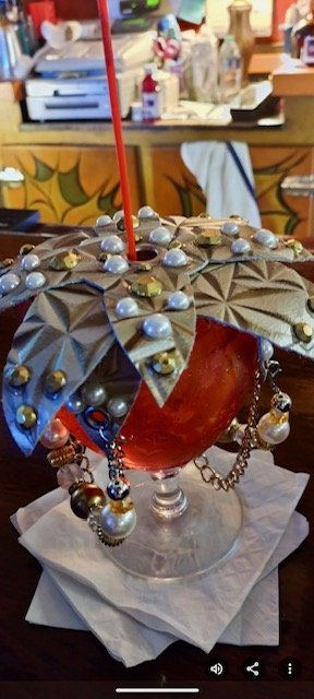 Decorative drink served in a large glass, topped with holiday-themed gold and pearl beads, stars, and artificial leaves, with a red straw, on a napkin, in a festive setting.