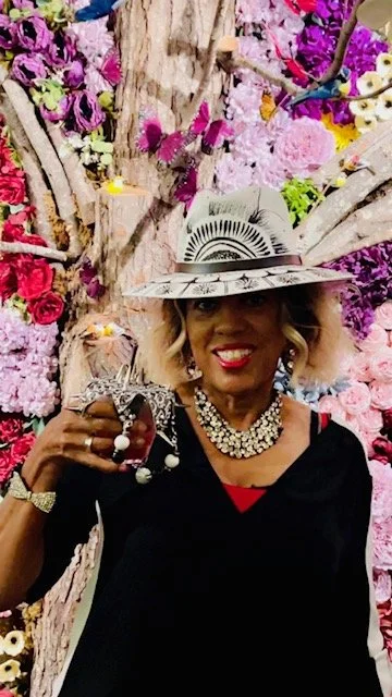 Woman smiling, wearing a stylish hat, holding a wine glass, standing in front of a colorful pink and purple floral backdrop.