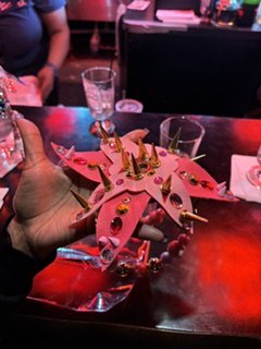 A person holding a pink unicorn-shaped cake with gold accents and small decorative elements, in a lively setting with drinks and tables.