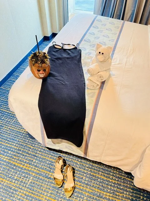 Halloween-themed pumpkin bucket, black dress, white rolled towel shaped like a bear, pair of high-heeled shoes, on a hotel bed with striped curtains in the background.