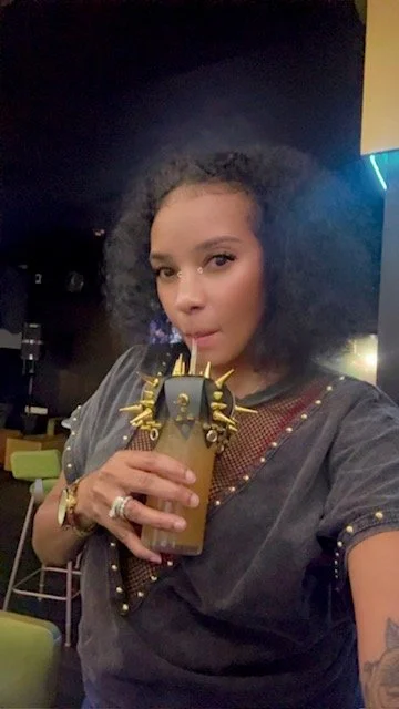 Woman with curly hair drinking a beverage from a tall cup with a yellow straw in a dimly lit environment.