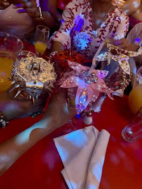 People celebrating at a party with colorful, decorated items on the table, including a pink unicorn-shaped object with sequins and rhinestones, surrounded by glasses and drinks, under vibrant lighting.