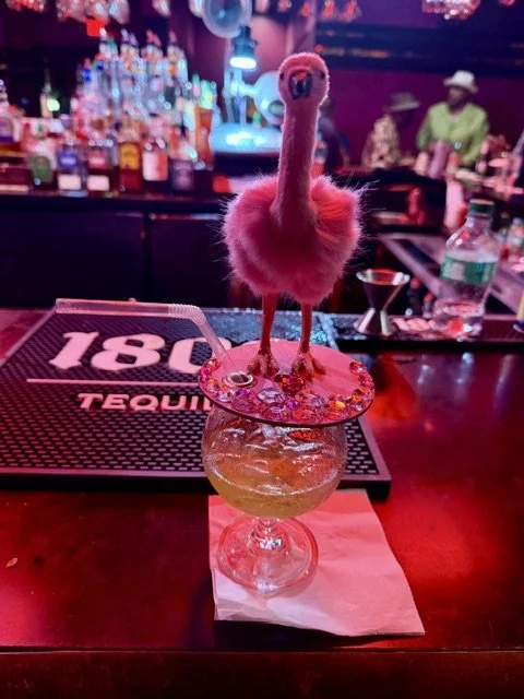 A glass on a bar counter holding a pink flamingo drink with a small plush flamingo on top, with bottles and bar patrons in the background.