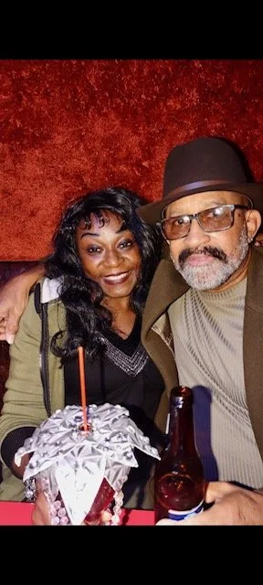 A man and woman sitting together at a table, smiling, with a birthday cake with a single candle and a bottle of beer visible in front of them. The background is red.