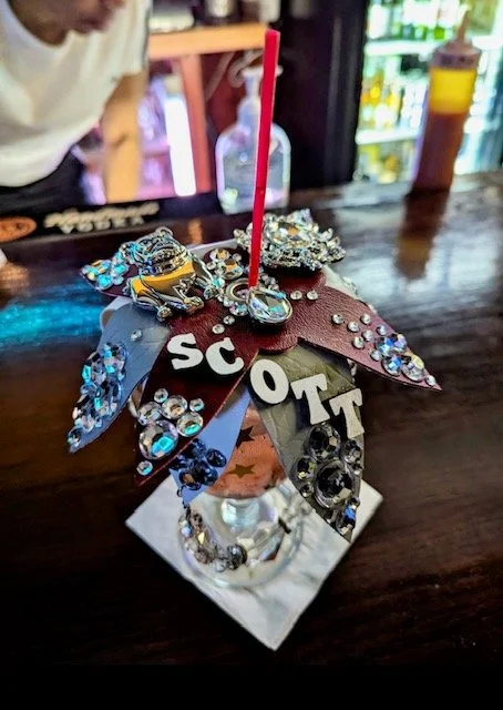 A decorated protest sign with the word 'SCOT' and various rhinestones and embellishments, attached to a silver stick in a glass container on a wooden table.