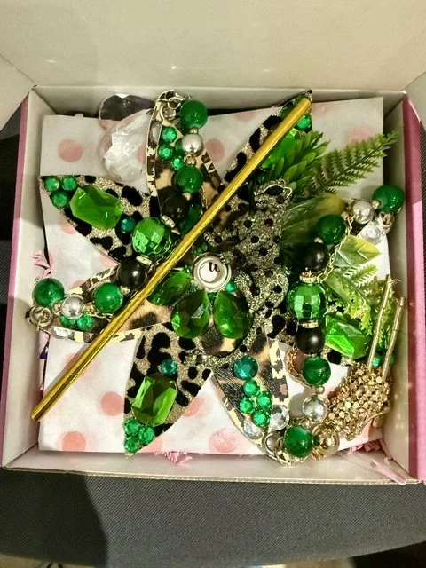 A decorative jewelry box containing green, silver, and black beaded jewelry, a gold hairpin, synthetic leaves, and a large black and white flower-shaped accessory with a leopard print design.