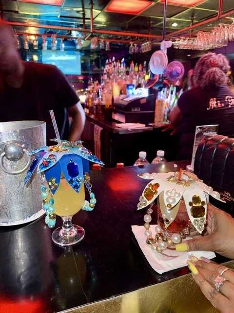 Two decorated drinks with small umbrellas and beads on a bar counter in a lively bar setting with people and colorful bottles in the background.