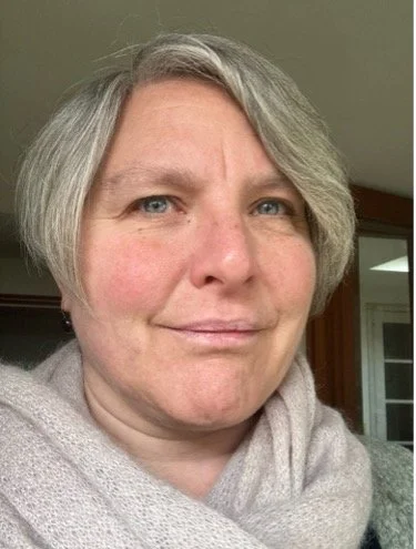 A white woman wearing a grey scarf smiles slightly at the camera.