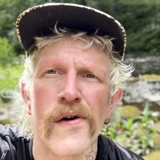 A light-skinned man with blond hair and a moustache wears a hat and looks at the camera.