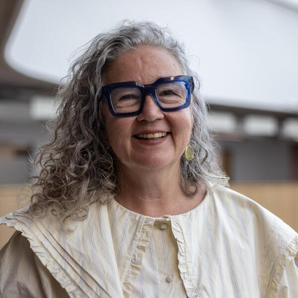 A woman with curly hair and thick-framed blue glasses smiles at the camera.