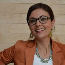A white woman with dark hair and dark-rimmed glasses smiled at the camera. She is wearing an orange jacket with a light blue necklace.