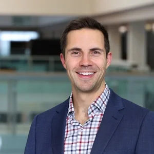 A white man in a plaid shirt with a jacket smiles at the camera.