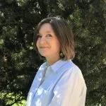 A white woman with short brown hair stands in front of foliage and slightly smiles at the camera.