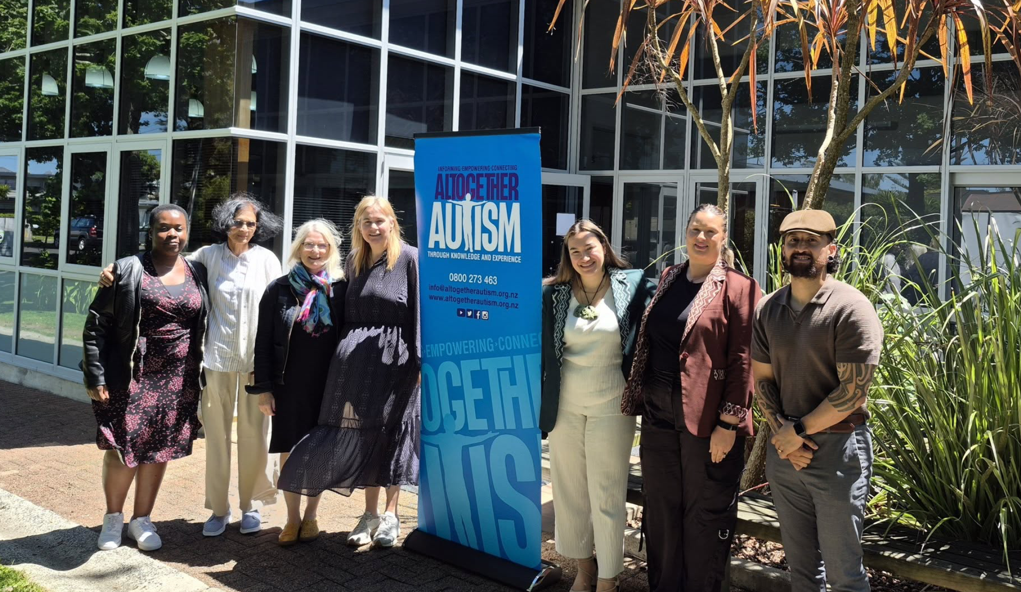 An image of seven Māori, Pacifika people and settlers standing shoulder to shoulder outside beside an Altogether Autism banner.