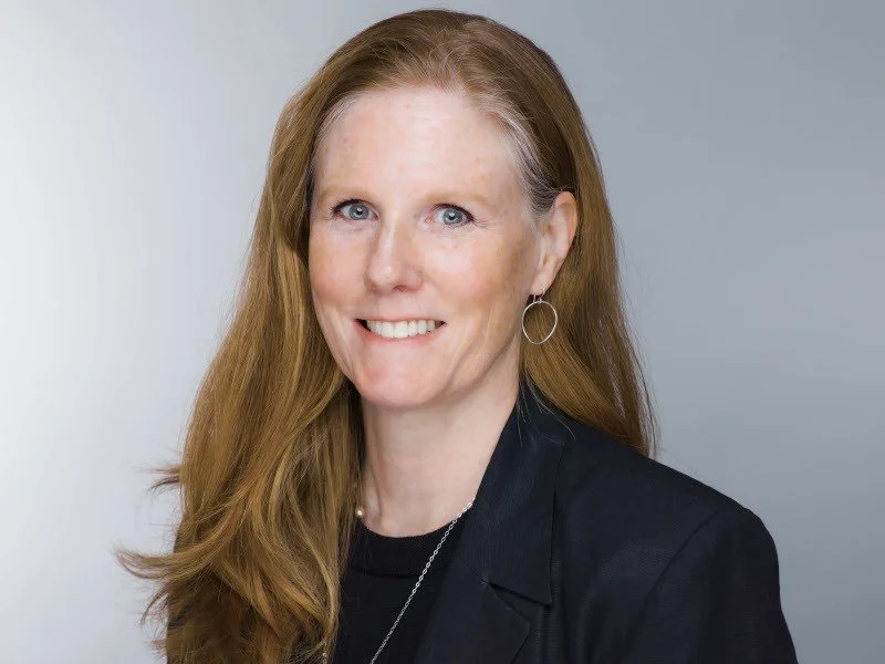 A light-skinned woman with reddish hair wears a black shirt and smiles at the camera.