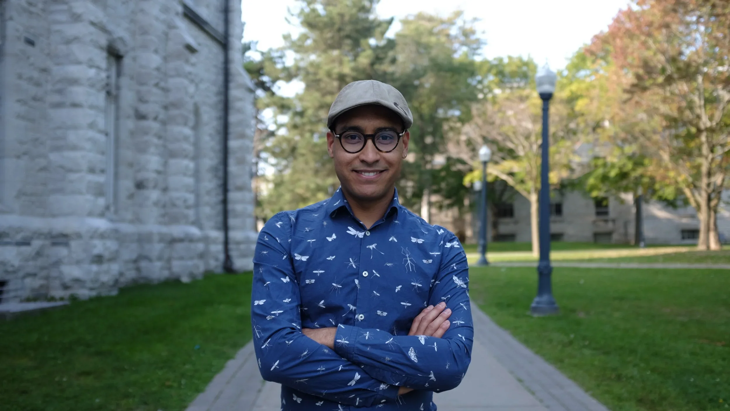 A man wearing a grey hat and round, dark-rimmed glasses crosses his arms and smiles at the camera.