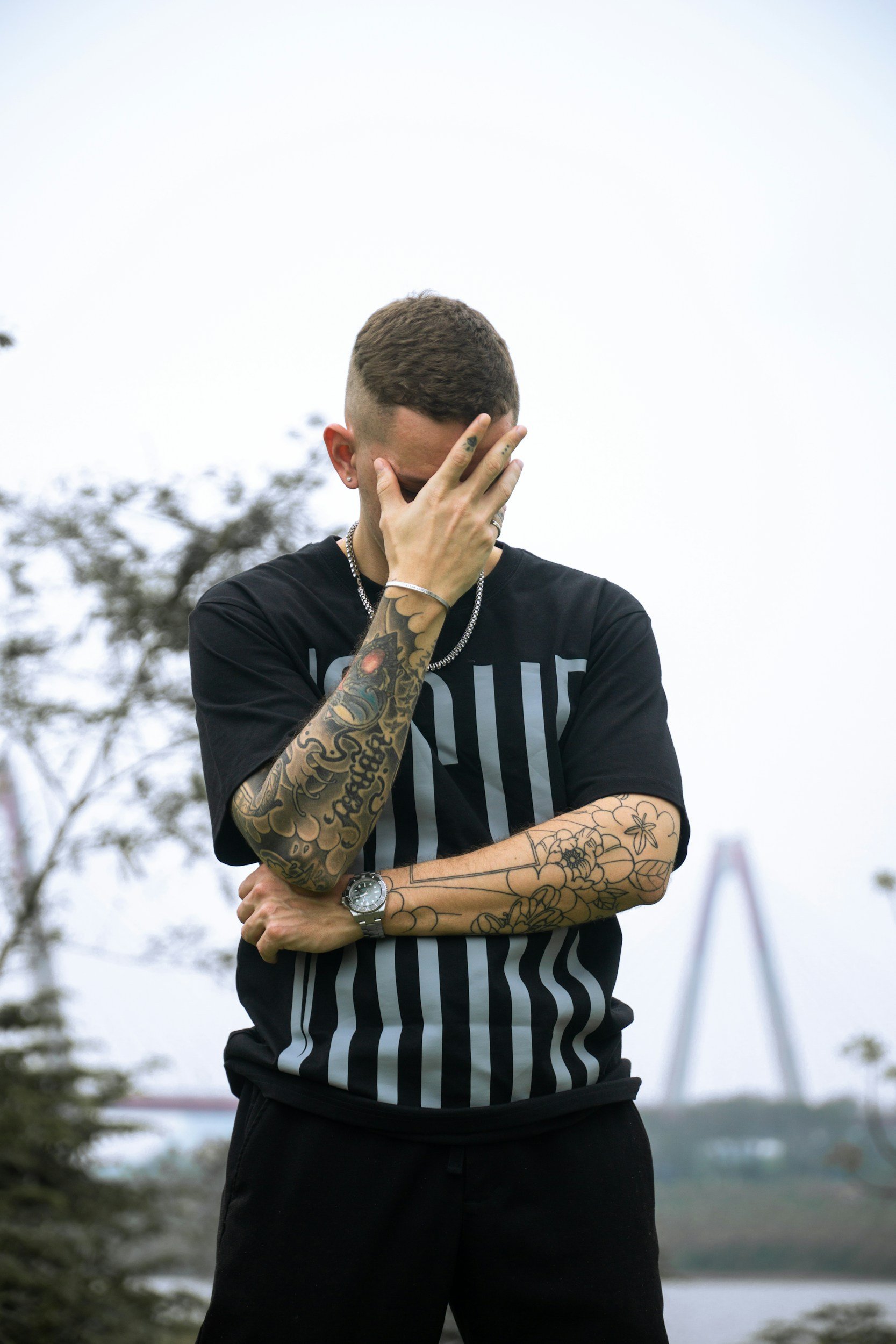 A man with tattoos on his arms and wearing a black T-shirt with vertical stripes is standing outdoors, covering his face with his hand. The background features trees and a bridge over a body of water.