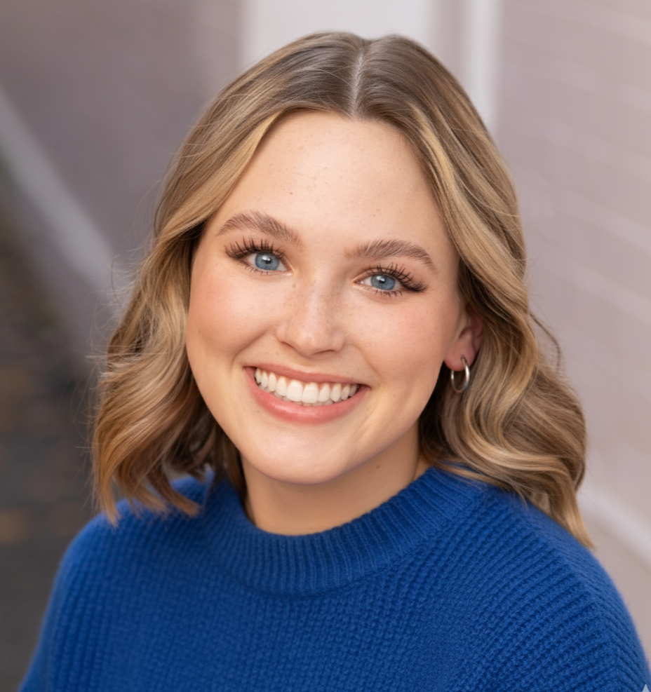 A young woman with short, wavy blonde hair, blue eyes, and a bright smile, wearing hoop earrings and a pink sleeveless turtleneck top, stands against a warm brown background.