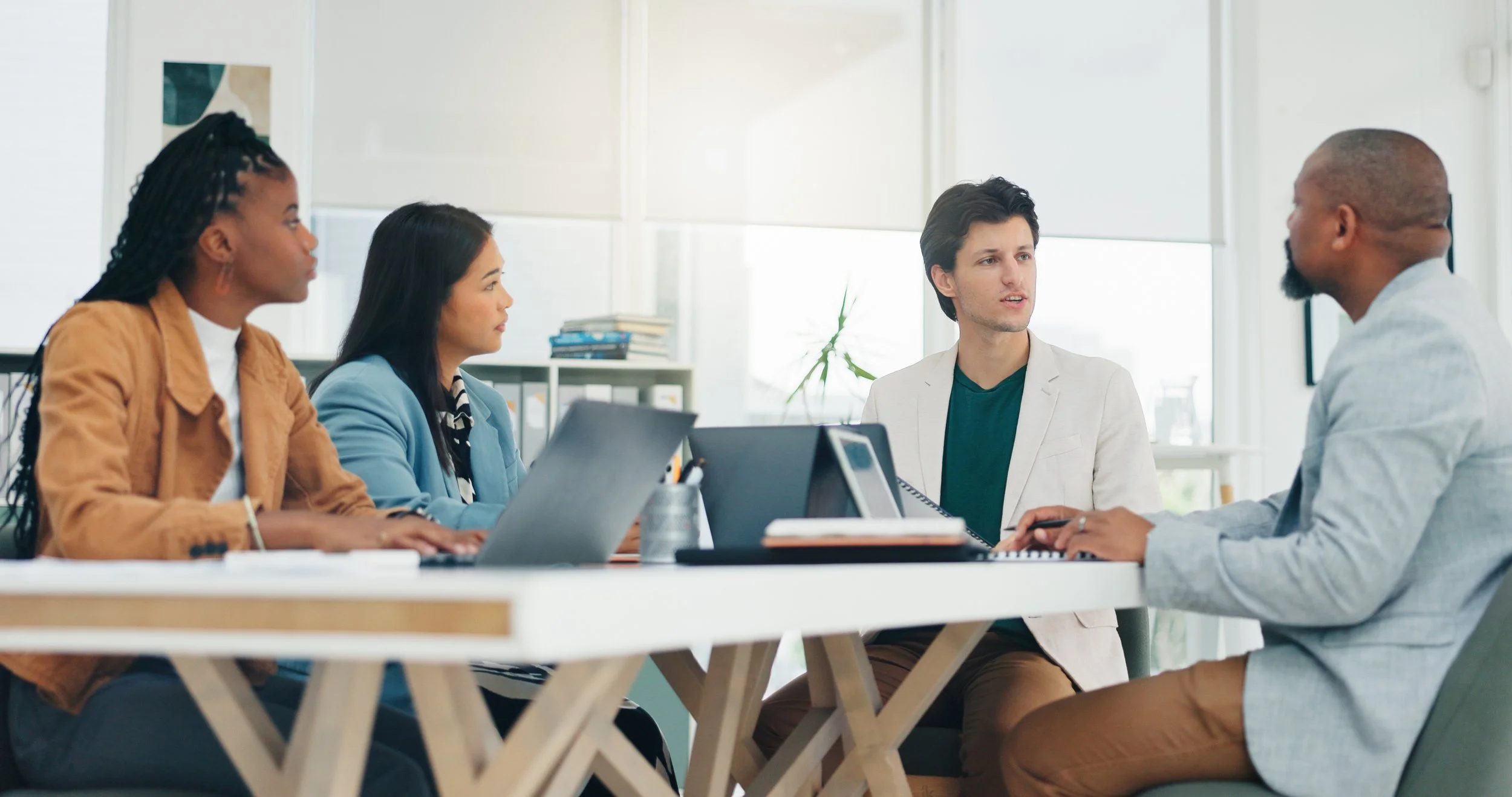 A diverse group of five young professionals, three women and two men, engaged in a meeting around a table with laptops and documents in a bright modern office.