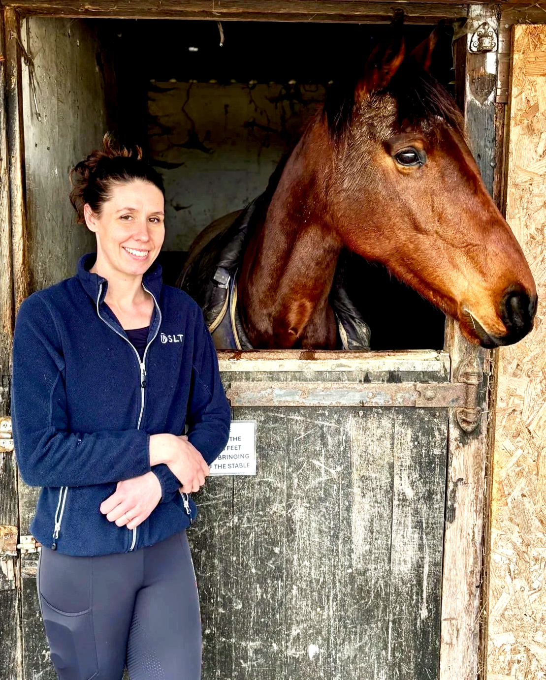 Woman smiling and leaning on a stable door, petting a white horse inside a barn.