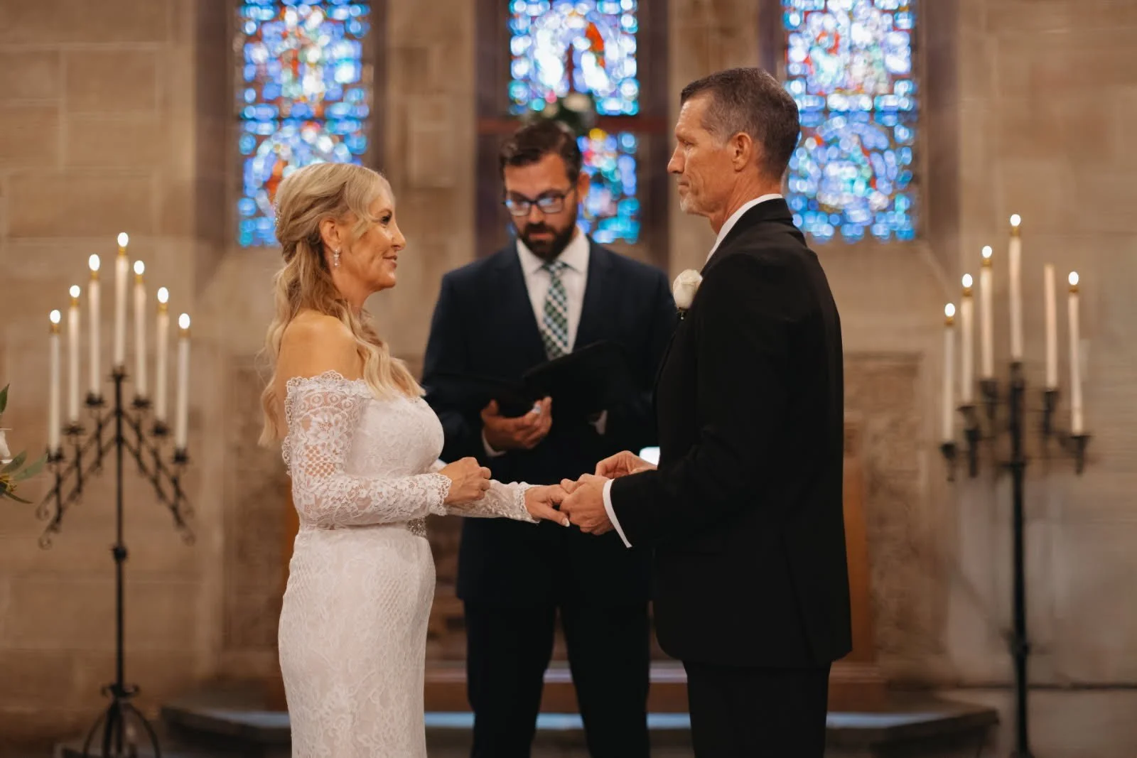 A bride and groom exchanging vows during a wedding ceremony inside a church, with a minister standing behind them, and stained glass windows in the background.