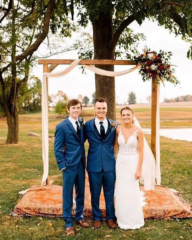 Wedding ceremony with a bride and two grooms standing under an outdoor wooden arch decorated with sheer fabric and flowers, set in a grassy field with trees and water in the background.