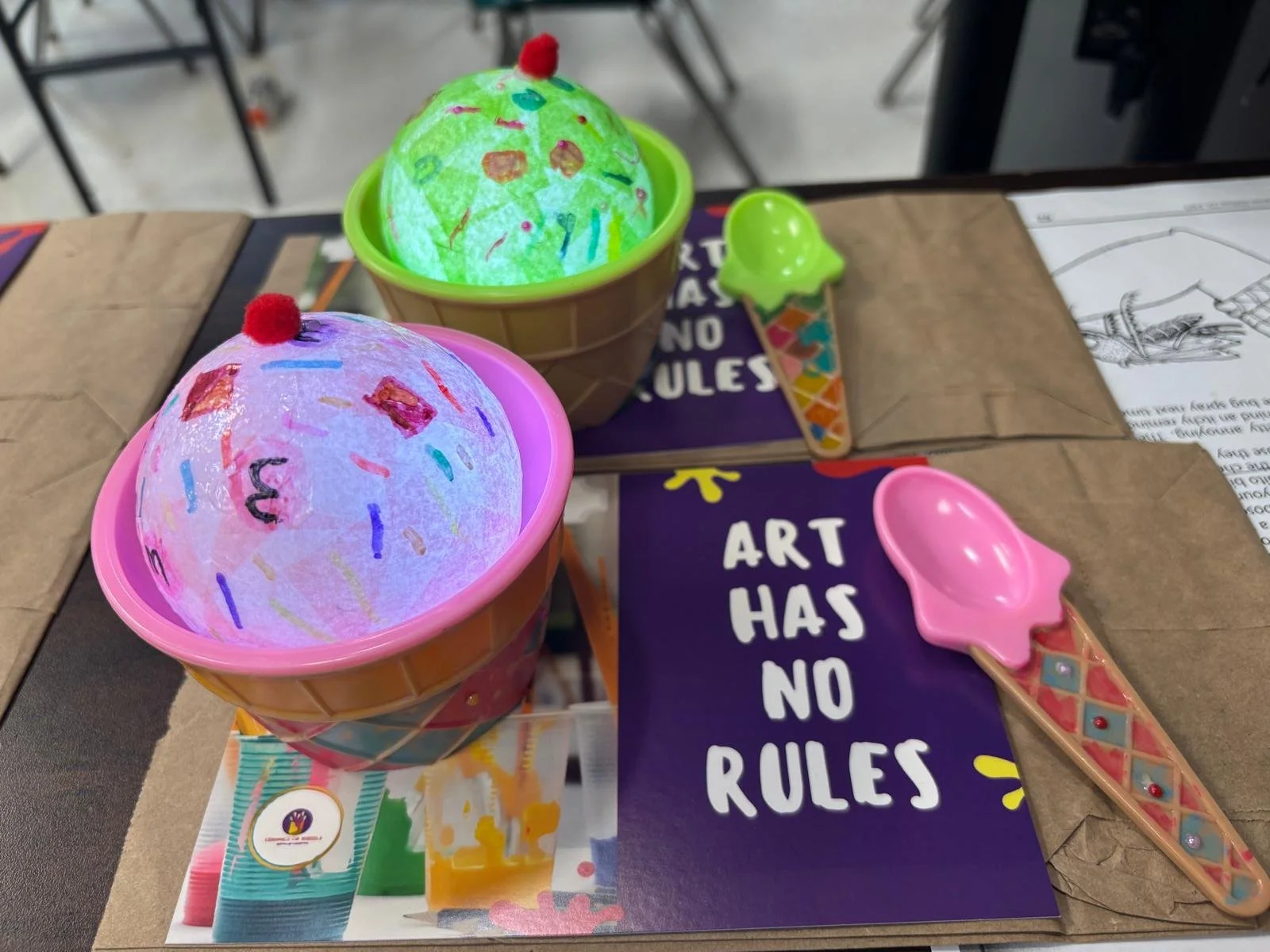 Two illuminated paper lanterns decorated like ice cream cones placed on top of books and papers on a table. The lanterns have colorful sprinkles and a small red pom-pom on top, resembling cherry toppings. Two plastic ice cream cone shapes are also on the table, one green and one pink, along with a purple sign that reads, 'ART HAS NO RULES.'