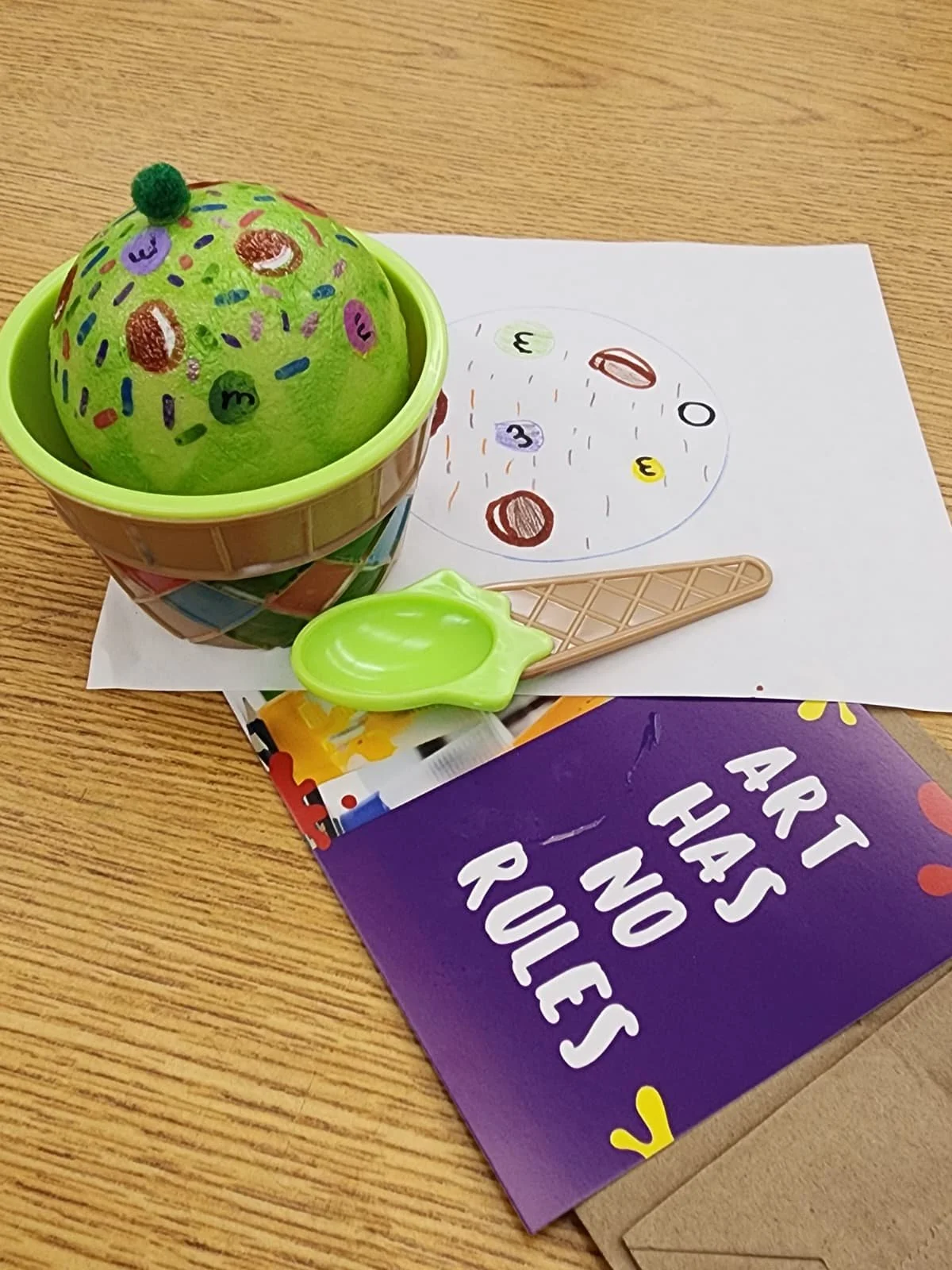 Ice cream-themed sensory art project created during a Ceramics on Wheels workshop, featuring a hand-painted green ice cream scoop with colorful candy-inspired designs, a toy cone, and activity materials.